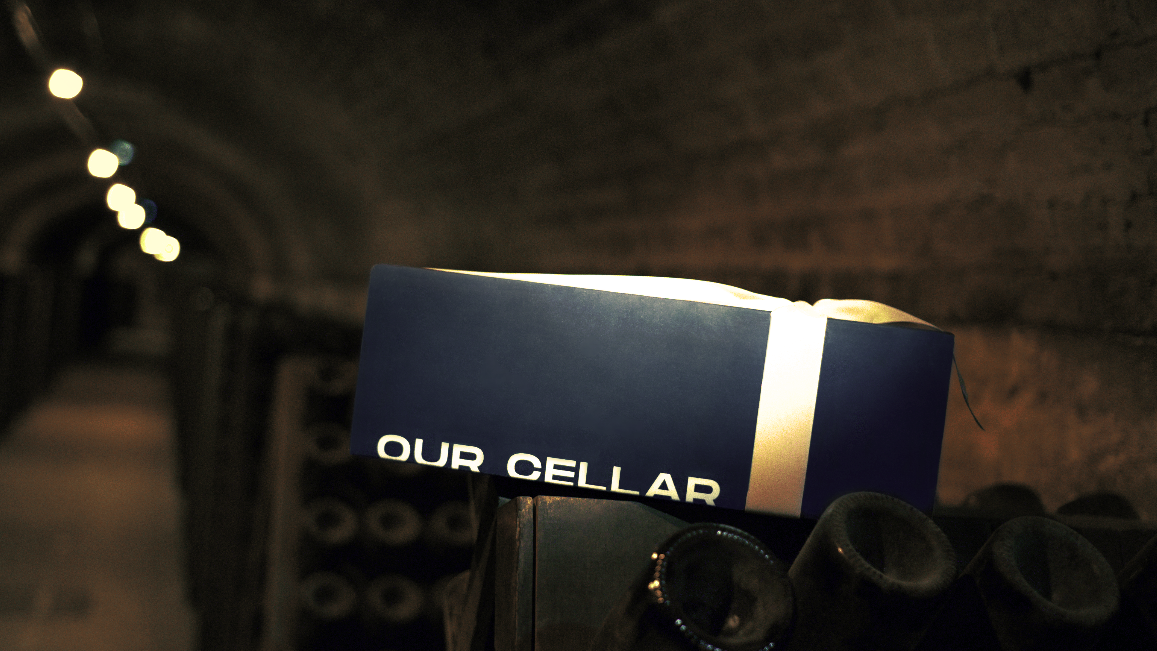 The box in a candlelit cellar, on top of bottles in repose. Shows the brand world without saying the word "luxury."